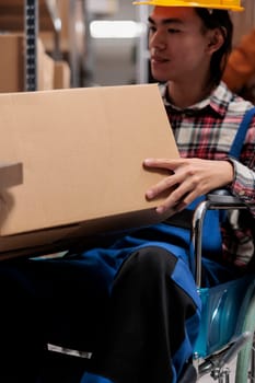 Asian warehouse order picker wheelchair user holding customer parcel. Young storehouse employee with disability preparing freight for shipment while working in inclusive workplace