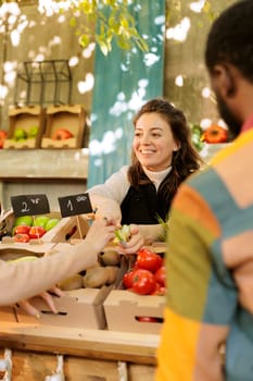 Smiling woman vendor offering customer to try out small piece of organic apple while selling fresh natural fruits and vegetables at harvest fair festival, selective focus. Tasting during shopping.