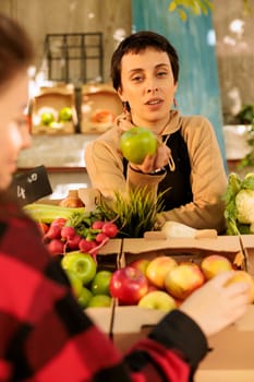 Local vendor wearing an apron and talking to customers while selling organic fresh apples at a farmers market stand. A young woman holds bio natural farming products grown locally.