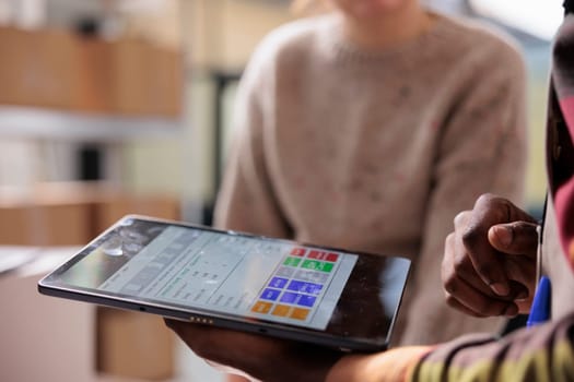 Stockroom managers analyzing merchandise report inventory on digital device, preparing clients orders before start shipping packages. Diverse team working in warehouse. Close up shot