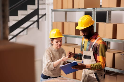 Diverse stockroom looking at products logistics report, while preparing clients orders in warehouse. African american supervisor working at products inventory with worker in storage room