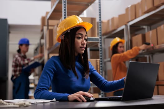 Asian retail storehouse employee analyzing orders checklist on laptop while working in storage room. Warehouse manager supervising inventory management while using computer