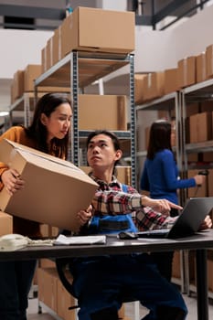 Warehouse asian workers holding parcel and scheduling delivery on laptop. Storehouse employees discussing orders checklist and analyzing pick ticket on computer in storage room