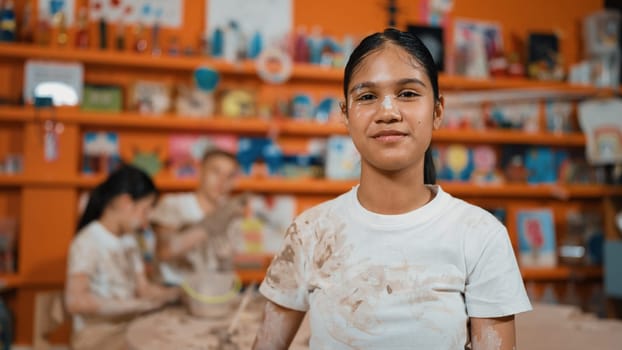 Smiling student looking at camera while standing at workshop while diverse student having pottery class together. Happy caucasian girl smile while wear dirty shirt in art lesson. Edification.