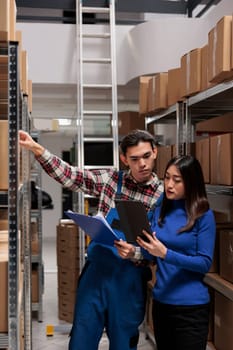 Warehouse employees managing inventory tracking with goods checklist. Storehouse asian man and woman workers picking order while standing in storage room aisle with shelves full of cardboard boxes
