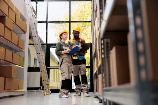 Diverse team wearing helmet and industrial overall, checking cardboard boxes for packages shipping in warehouse. Storage room employess preparing customers orders while doing goods quality control