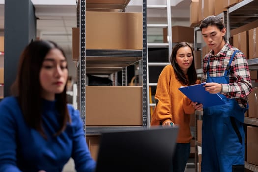 Delivery managers analyzing customer orders list clipboard in ecommerce retail business warehouse. Storehouse asian man and woman employees checking inventory report data in storage room