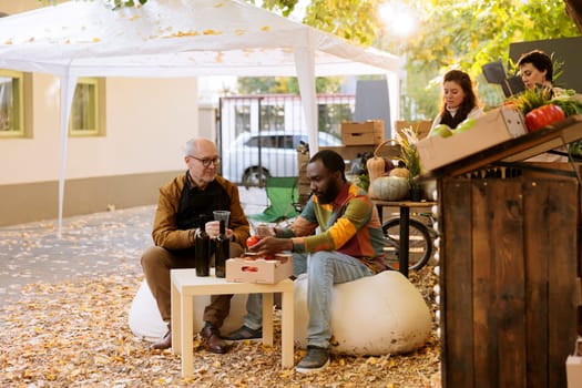 African american customer sitting at a farmers market table with a local vendor, having homemade wine. Black male client visiting an organic autumn festival and enjoying wine and food tasting.