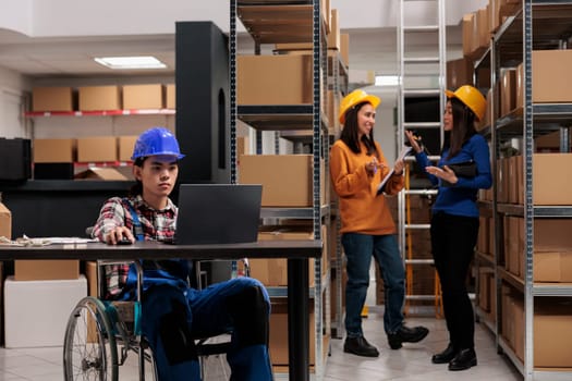 Warehouse worker in wheelchair planning delivery on laptop in logistics and distribution center storage room. Young asian storehouse employee with disability analyzing clients orders checklist