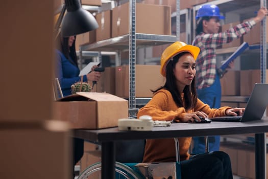 Asian postal service worker with physical disability using laptop in warehouse. Young woman wearing yellow protective hardhat in wheelchair tracking parcels shipment online on computer