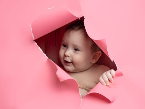 Cute Caucasian newborn baby boy peeks out of a hole in a paper pink background