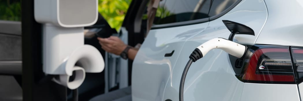 Young man recharge electric car's battery from charging station in outdoor green city park. Rechargeable EV car for sustainable environmental friendly urban travel. Panorama Expedient