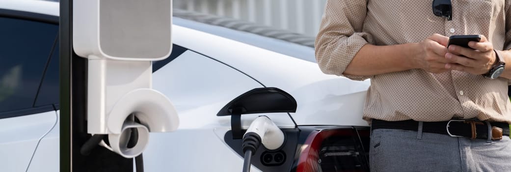 Young man use smartphone to pay for electricity at public EV car charging station at city commercial mall parking lot. Modern environmental and sustainable urban lifestyle with EV vehicle. Expedient