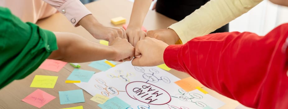 Young happy creative startup group join circle fist bump hands together surrounded by marketing strategy mind map and colorful sticky notes at meeting room. Unity and teamwork concept. Variegated.