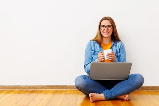 Happy woman sitting on floor with laptop and coffee, casual lifestyle. Home office concept with copy space for design and print.