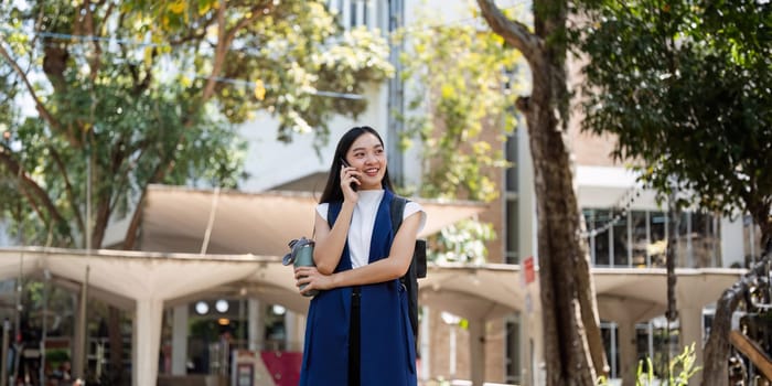 Young cheerful Asian successful businesswoman standing on city street talking on mobile phone.