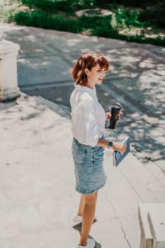 Business woman city street. A woman in a white shirt and blue denim skirt walks down the street holding a blue notebook in her hands. Summer city walks, travel.