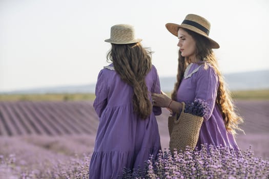 Mom and daughter are running through a lavender field dressed in purple dresses, long hair flowing and wearing hats. The field is full of purple flowers and the sky is clear