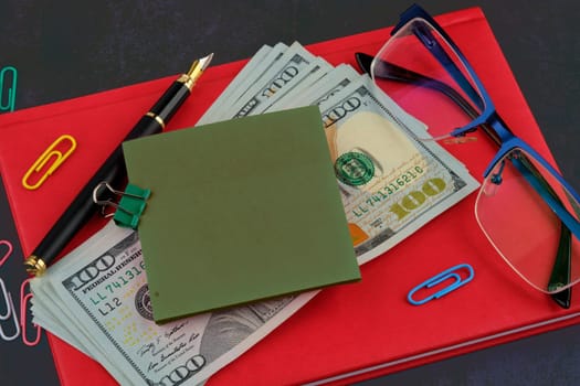 A sticker for writing attached to dollars on a red business notebook, glasses next to it, a pen on a dark background