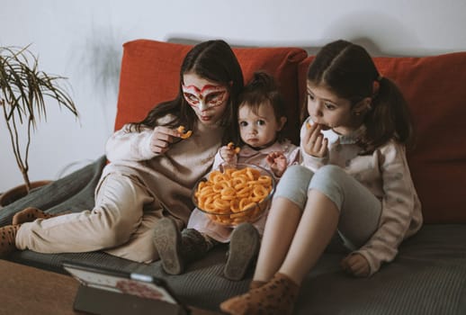 Three beautiful caucasian girls sisters are sitting on the sofa,eating corn rings and emotionally watching a movie on the tablet in the room in the evening,close-up side view.The concept of family watching movies.