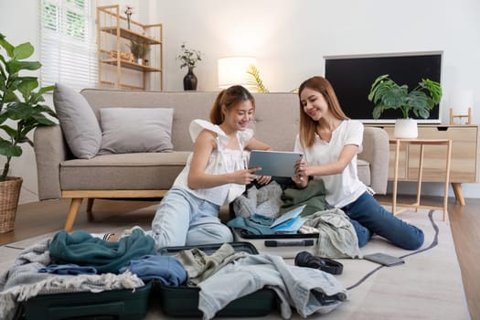 Young Asian woman packing clothes to the suitcase. Preparation for the summer vacation. Two women are planning and with using tablet for trip booking a trip and helping to prepare luggage to travel.