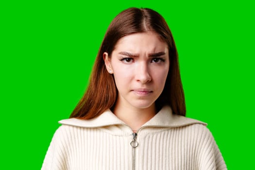 A young woman is captured against a vivid green backdrop. She is wearing a white zip-up top and has her brown hair styled straight. The womans face is showing a confused or perplexed expression, with her eyebrows furrowed as if shes trying to understand something or is in slight disbelief.