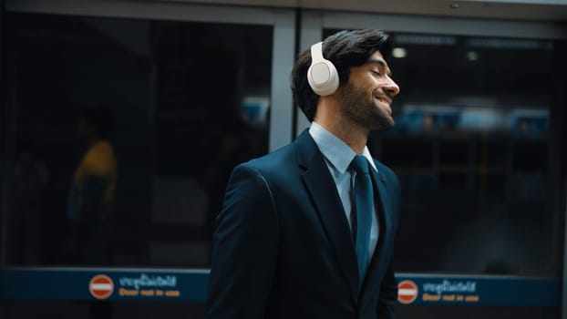Closeup of smart executive manager listen relaxing music while waiting at train station. Happy male leader wear headphone and standing at door area. Public transport concept. Dark filter. Exultant.