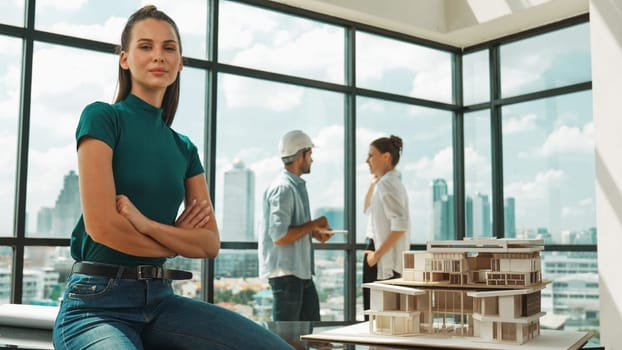 Young beautiful interior designer crossing arms while engineer team talking about house design. Skilled businesswoman smiling at camera with confident while sitting near house model. Tracery