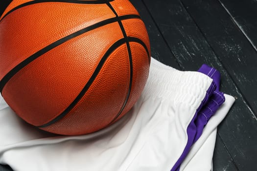 A freshly polished orange basketball sits on a dark wooden surface, contrasting with the deep black background. A white uniform with purple trim is casually draped next to the ball, suggesting a pause in play or the aftermath of a game.