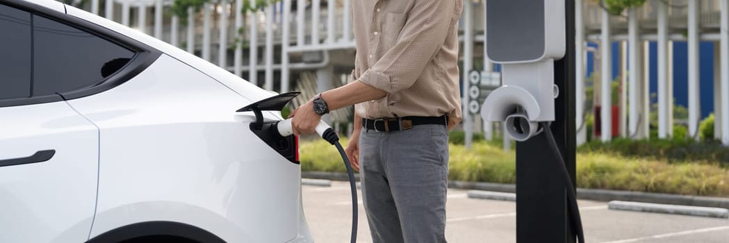 Young man recharge electric car's battery from charging station in city commercial parking lot. Rechargeable EV car for sustainable environmental friendly urban travel. Panorama Expedient