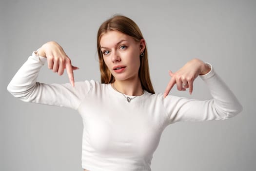 Young Woman in White Shirt Pointing Downwards With Both Hands Against a Gray Background in studio