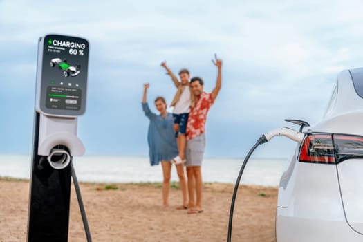 Alternative family vacation trip traveling by the beach with electric car recharging battery from EV charging station with blurred cheerful and happy family enjoying the seascape background. Perpetual