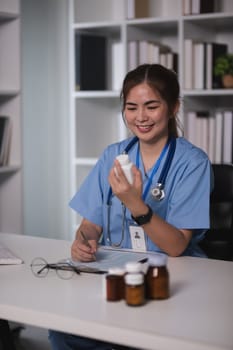 A woman in a blue scrubs is smiling as she holds a bottle of pills. She is sitting at a desk with a clipboard and a keyboard. The scene suggests a friendly and positive atmosphere