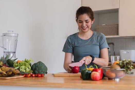 A woman is cutting an apple on a wooden cutting board in a kitchen. She is smiling and she is enjoying the process. The kitchen is well-stocked with fresh fruits and vegetables, including apples