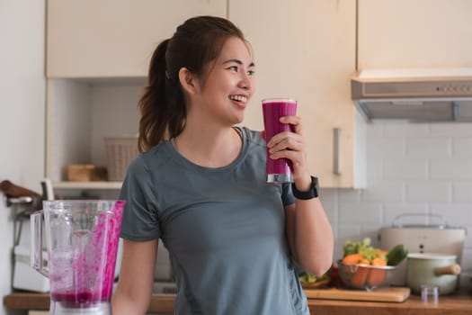 A woman is smiling while holding a pink drink in a blender. She is in a kitchen with a counter full of food and utensils