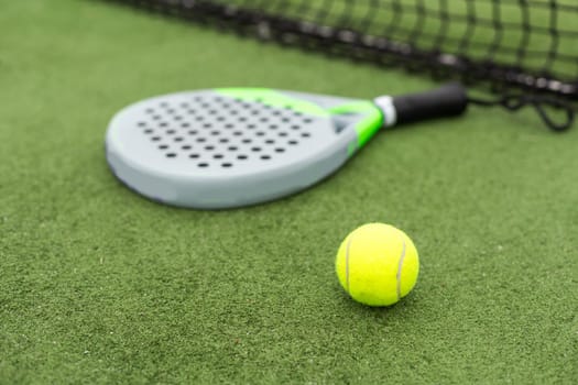 Yellow balls on grass turf near padel tennis racket behind net in green court outdoors with natural lighting. Paddle is a racquet game. Professional sport concept with copy space. High quality photo