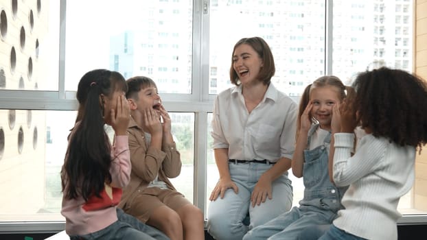 Smart caucasian teacher teaching while sitting on floor together surrounded by diverse students. Professional instructor playing with multicultural learner. Creative education concept. Erudition.