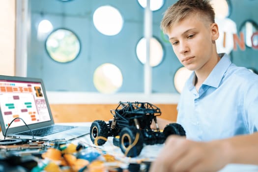 Schoolboy watching and considering parts of robotics car while learning technology in STEM class. On table put laptop, controller, electric wire, battery charger, and robotic vehicle. Edification.