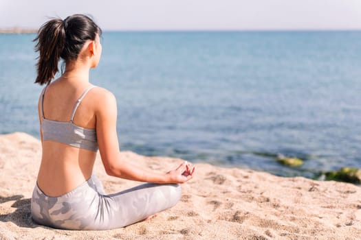 unrecognizable young woman in sportswear doing meditation at beach sitting with legs crossed, concept of mental relaxation and healthy lifestyle