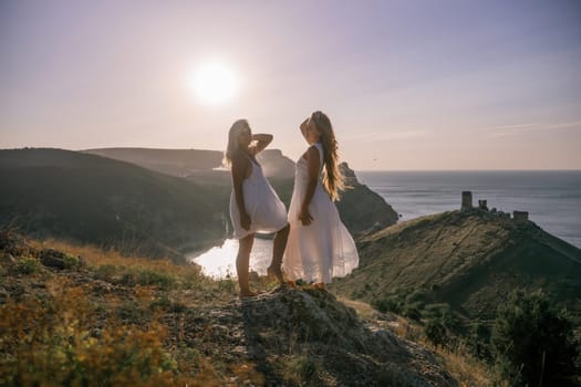 Two young girls are standing on a hillside, one of them wearing a white dress. The sun is shining brightly, creating a warm and inviting atmosphere. The girls seem to be enjoying their time together