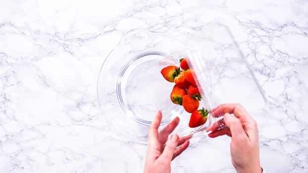 Flat lay. Ripe strawberries are submerged in water within a large glass mixing bowl, a step in washing the fruit to ensure cleanliness and longevity before storage or consumption.