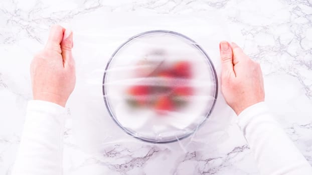 Flat lay. Strawberries, freshly washed and dried, are neatly stored in a glass bowl lined with a paper towel and securely covered with plastic wrap to maintain freshness.