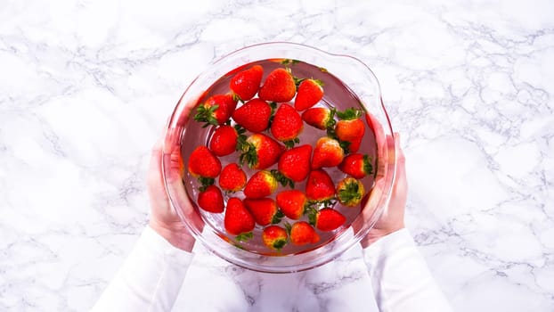 Flat lay. Ripe strawberries are submerged in water within a large glass mixing bowl, a step in washing the fruit to ensure cleanliness and longevity before storage or consumption.