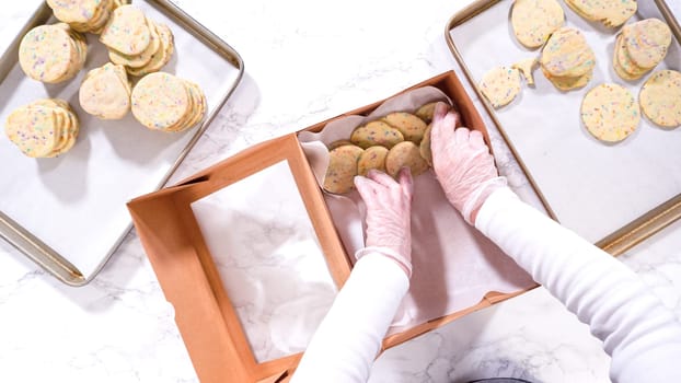 Flat lay. With precision, the woman is carefully arranging the sugar cookies, filled with dough-mixed sprinkles, into a rustic brown paper box.