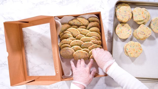 Flat lay. With precision, the woman is carefully arranging the sugar cookies, filled with dough-mixed sprinkles, into a rustic brown paper box.