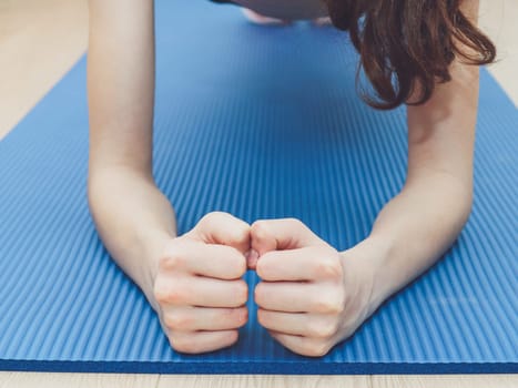 A young caucasian girl teenager without a face does a plank exercise with her hands clenched into fists on a blue mat, close-up side view. The concept of home fitness and healthy lifestyle.