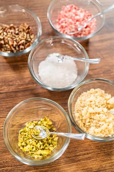 Ready-to-use cookie decorating supplies, including chopped nuts, colorful sprinkles, coarse sea salt, and golden toasted coconut, arranged in clear bowls on a wooden table.