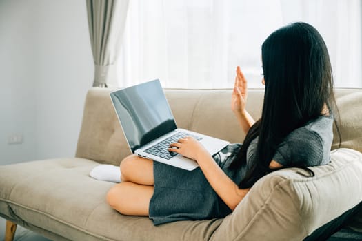 Woman on sofa uses laptop for video conferencing greeting friend. Engaged in distant meeting discussing laughing and learning. Modern online communication.