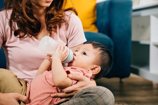 In the heart of their cozy home a mother smiles with joy as she feeds her cute Asian baby from a baby bottle in the living room capturing the pure essence of family togetherness.