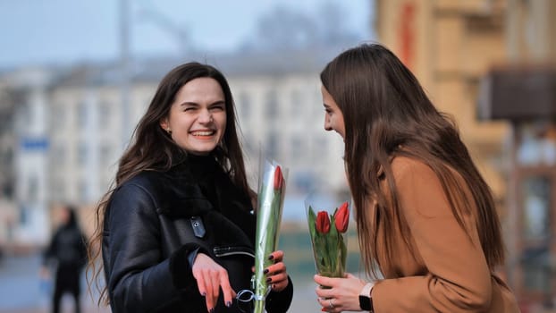 two happy girls, students, friends with flowers in their hands, laughing on a city street